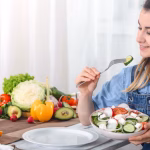 young-happy-woman-eating-salad-with-organic-vegetables-table-light-background-denim-clothes-concept-healthy-home-made-food